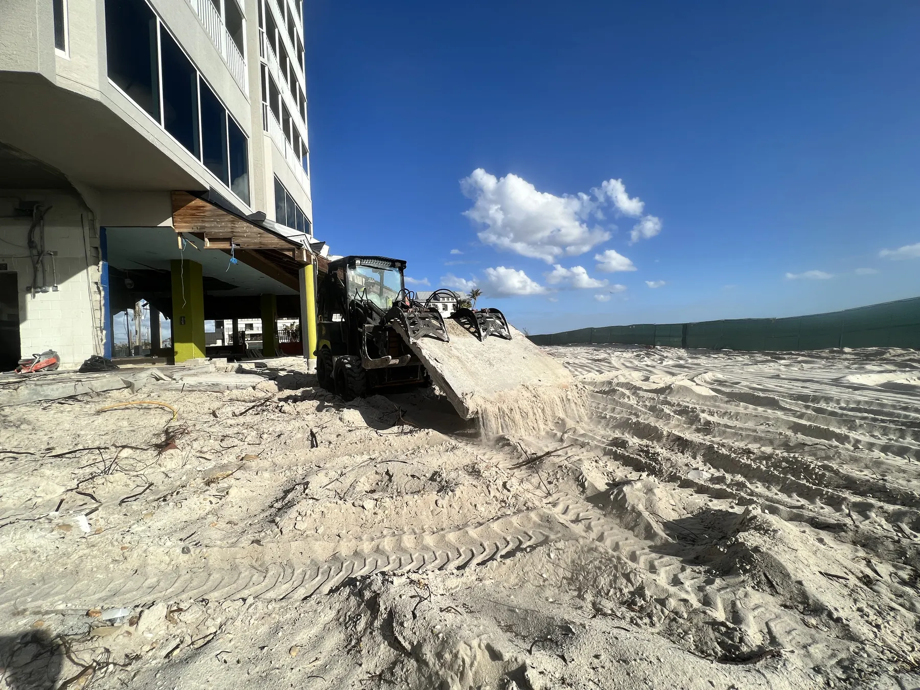 Tracked equipment grading sand beside a beachfront building and Gulf water