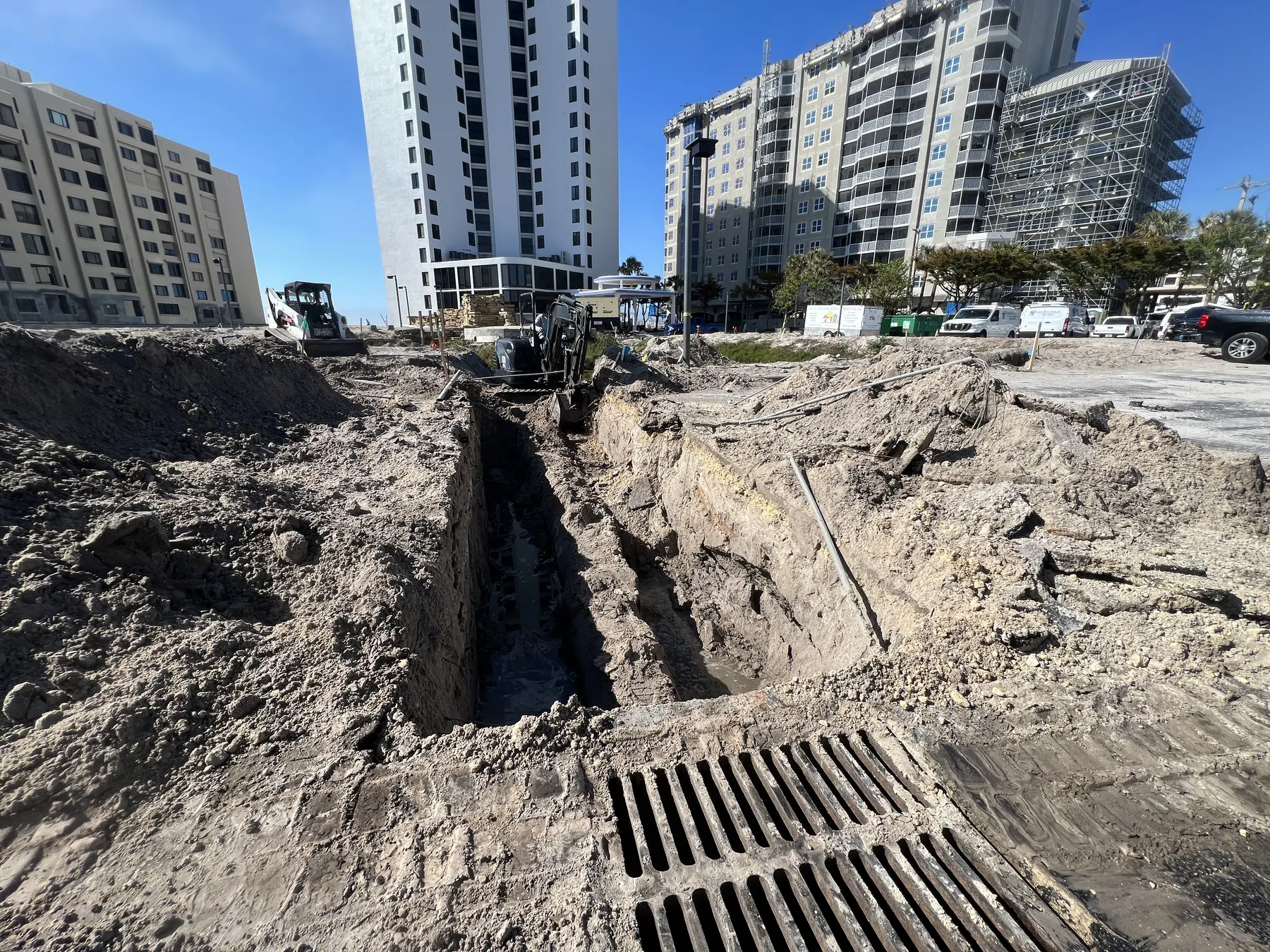 Deep drainage trench and grate work on a coastal construction site near high-rise buildings